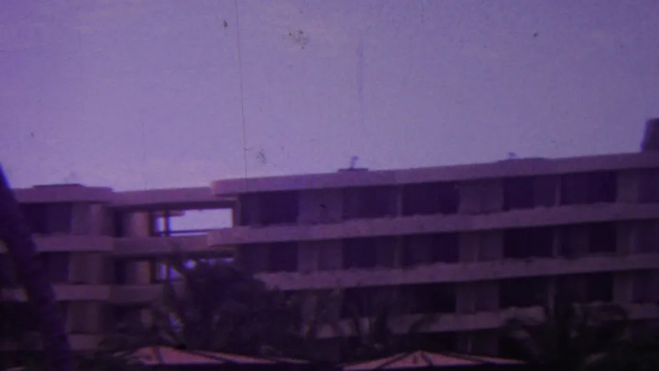 1962 Hawaii 8mm view of a multi-story hotel or motel exterior with horizontal balcony bands, palm trees, and dark window openings.