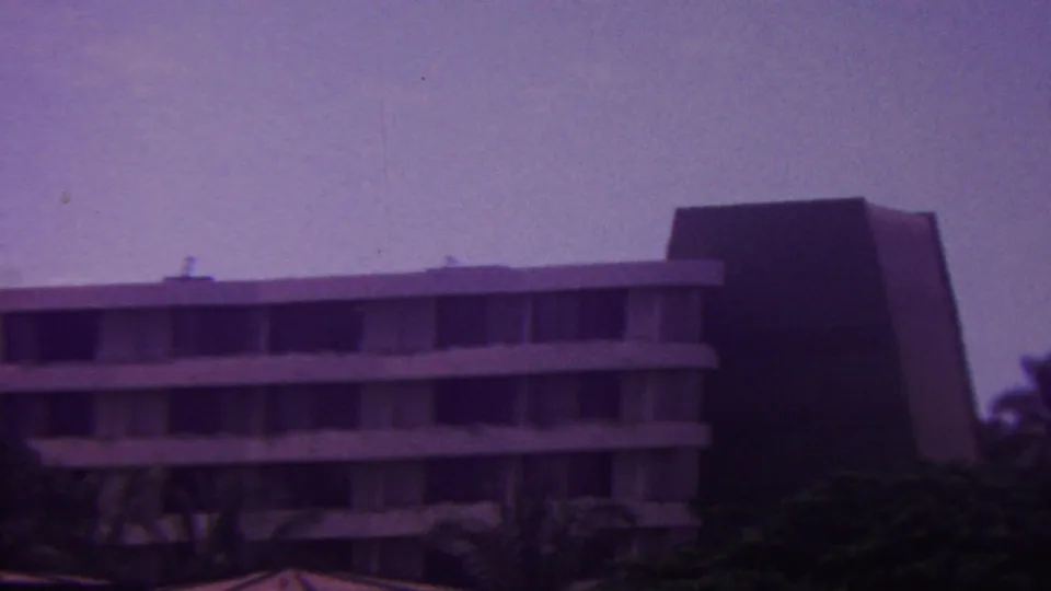 1962 Hawaii home-movie frame showing a resort-style hotel facade, palm trees, and a taller adjoining building at the right side of the composition.