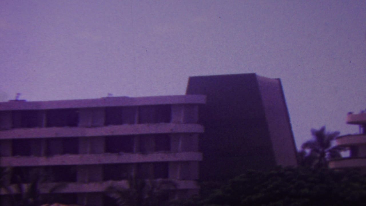 1962 Hawaii 8mm frame showing distant multi-story modern hotel buildings with flat balcony lines, dark angular walls, palms, and dense greenery.