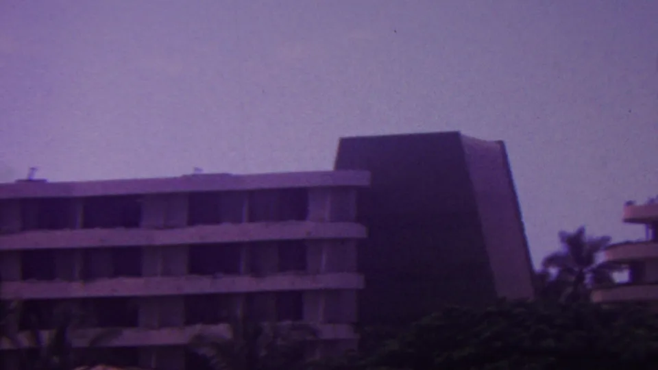1962 Hawaii 8mm frame showing distant multi-story modern hotel buildings with flat balcony lines, dark angular walls, palms, and dense greenery.