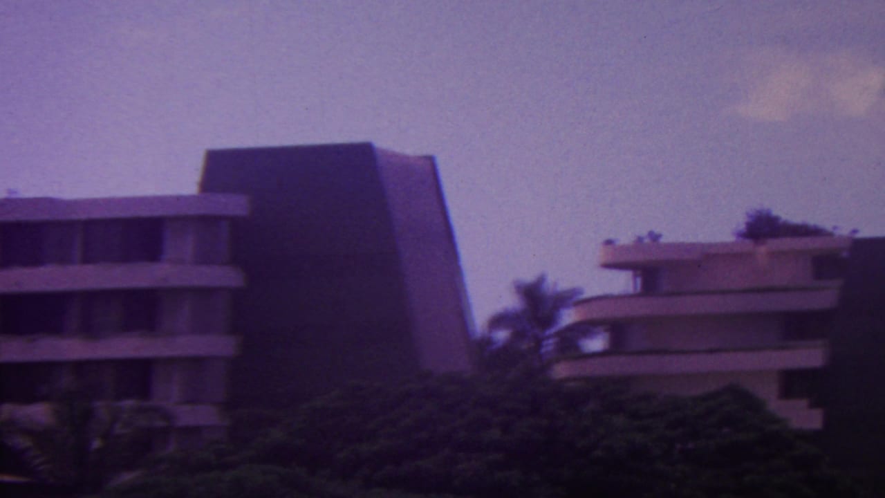 1962 Hawaii frame with a central modern hotel block, stacked balconies, dark side wall, palms, and soft cloud cover.