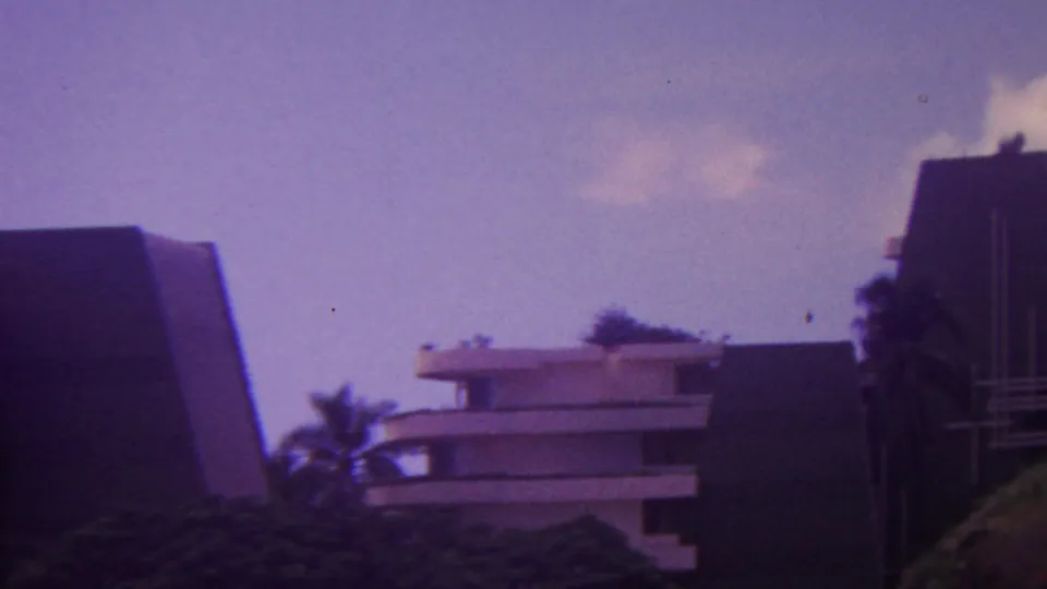 1962 Hawaii 8mm still showing hotel rooflines, a dark angular building face, palm foliage, and a soft purple film cast.