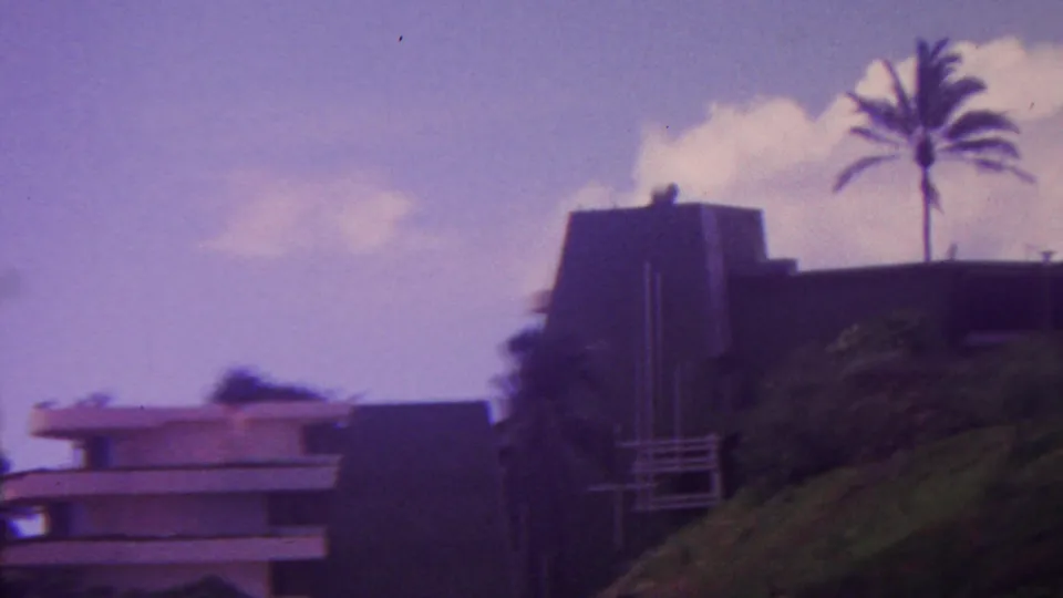 1962 Hawaii frame with a modern hotel section beside a dark wall, hillside greenery in front, and a tall palm rising against a cloudy sky.