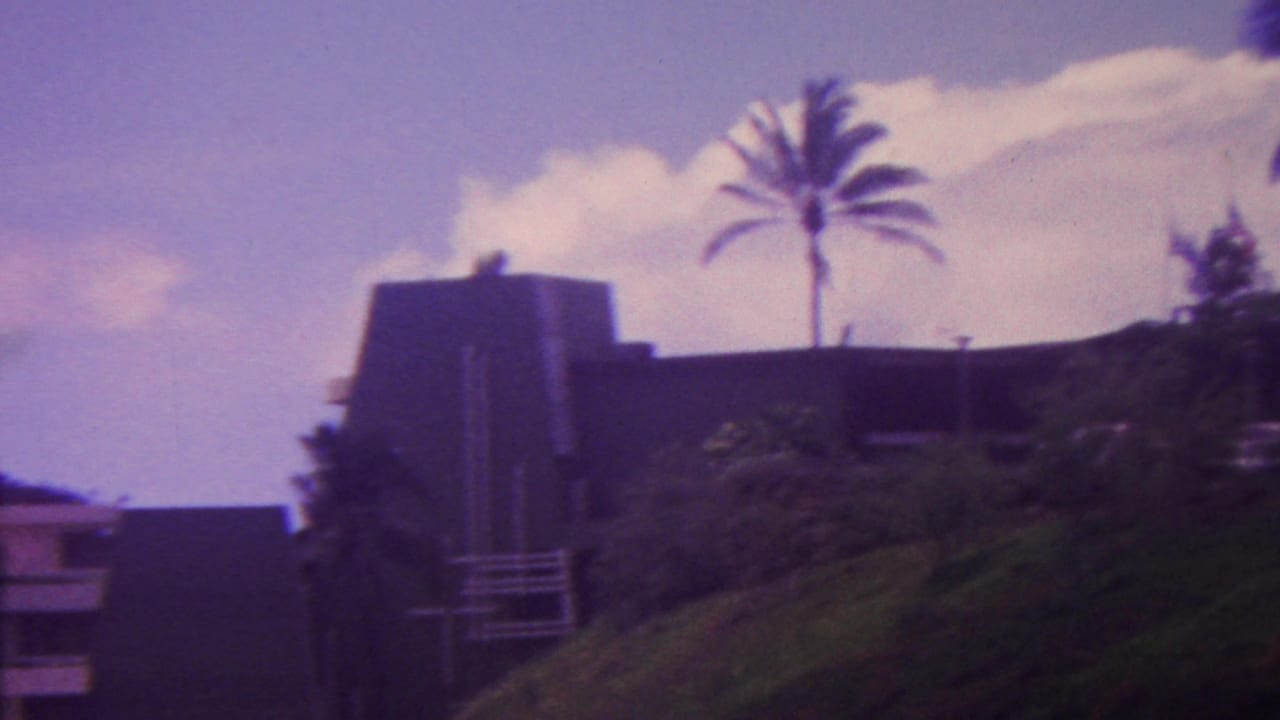 1962 Hawaii 8mm frame showing a dark hillside residence partly obscured by tropical vegetation, with tall palm trees and bright clouds overhead.