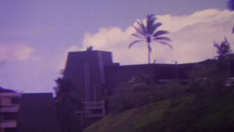 1962 Hawaii 8mm frame showing a dark hillside residence partly obscured by tropical vegetation, with tall palm trees and bright clouds overhead.