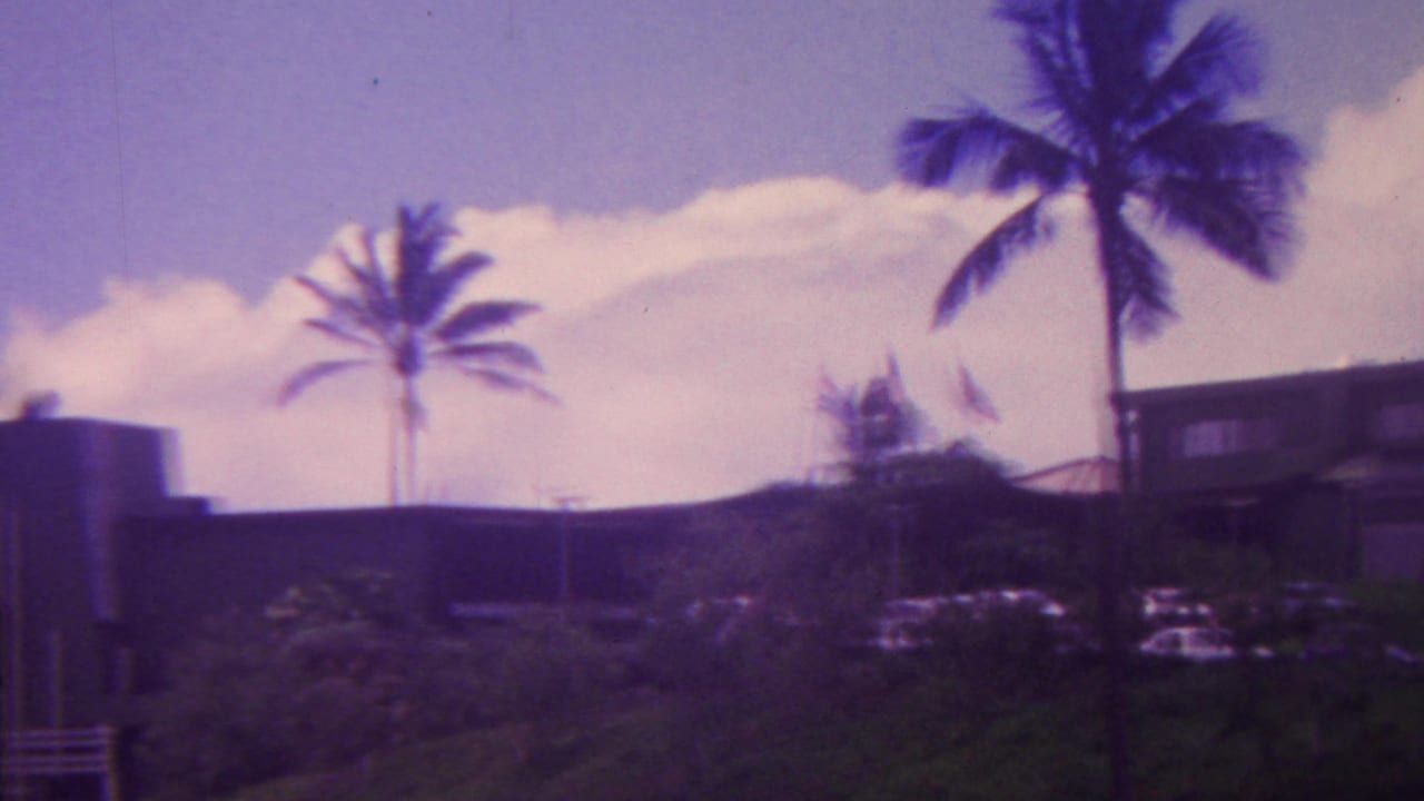 1962 Hawaii home-movie frame with a hillside residence, multiple palms, parked cars, and dense tropical landscaping under a bright cloud bank.