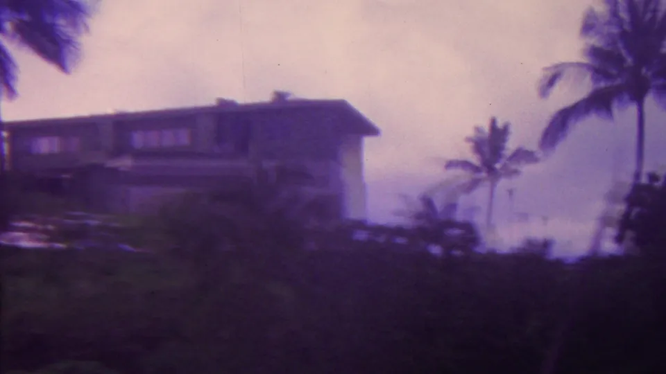 1962 Hawaii home-movie frame showing the residence from farther along the pan, with palms, dark vegetation, and a hazy distant landscape beyond the hillside.