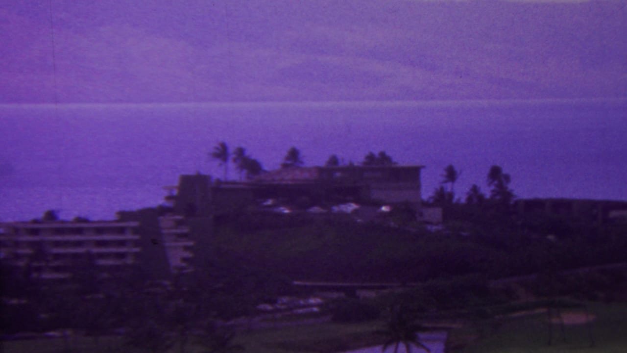 1962 Hawaii home-movie frame showing resort buildings on a hillside with palms, dark vegetation, and a broad ocean view beyond.