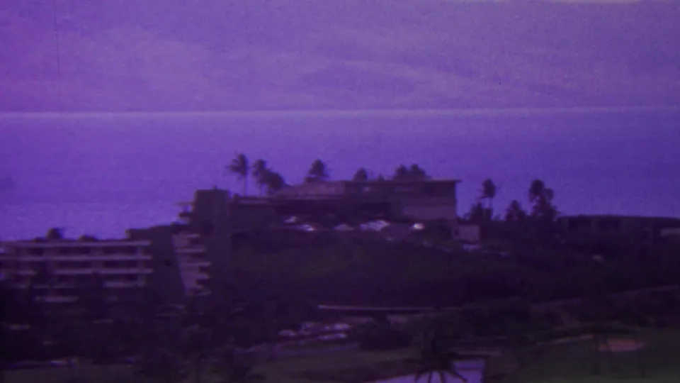 1962 Hawaii home-movie frame showing resort buildings on a hillside with palms, dark vegetation, and a broad ocean view beyond.