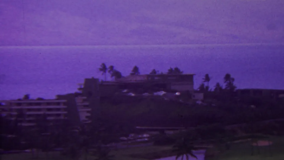 1962 Hawaii home-movie frame with an elevated view across resort structures, palm trees, dark landscaping, and the ocean horizon.