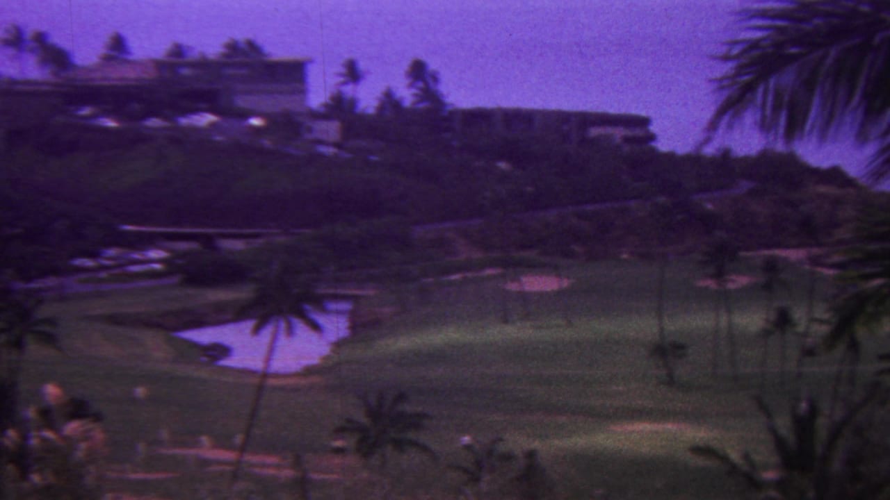 1962 Hawaii 8mm frame showing hillside resort buildings overlooking a green golf course with a pond, palms, and foreground vegetation under a purple film cast.