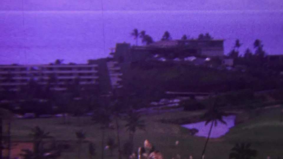 1962 Hawaii 8mm frame showing resort hotel blocks beside a landscaped golf course, pond, palms, and distant coastal haze.