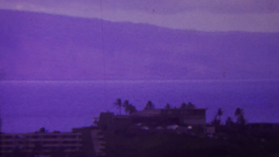 1962 Hawaii coastal resort view with distant ocean or bay, hazy mountain shapes, low hotel buildings, and palms in strongly purple 8mm footage.