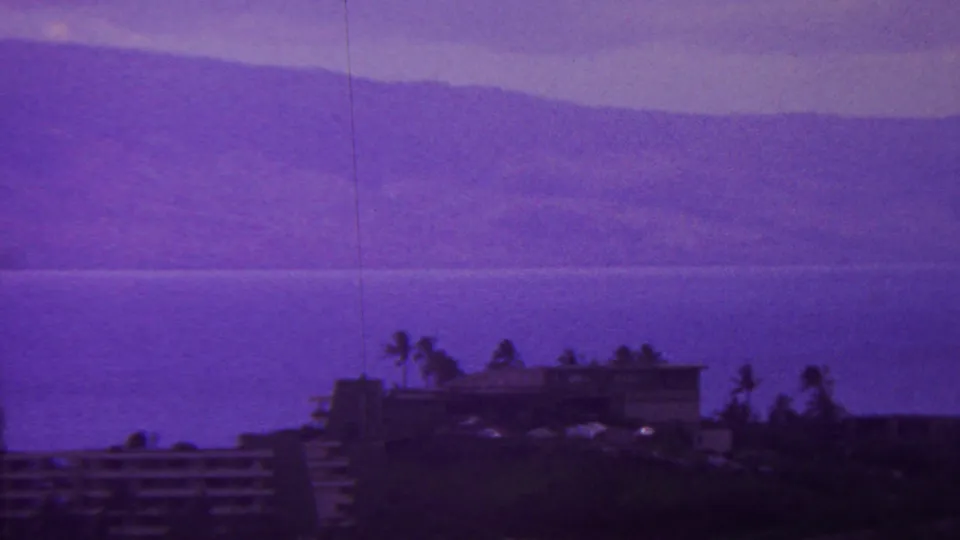 1962 Hawaii 8mm frame showing a distant shoreline building with palm silhouettes, ocean water, hazy mountains, and a purple-shifted sky.