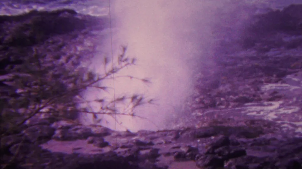 1962 Hawaii 8mm frame of a coastal blowhole-like spray plume rising from dark rocks as surf moves across the shoreline.