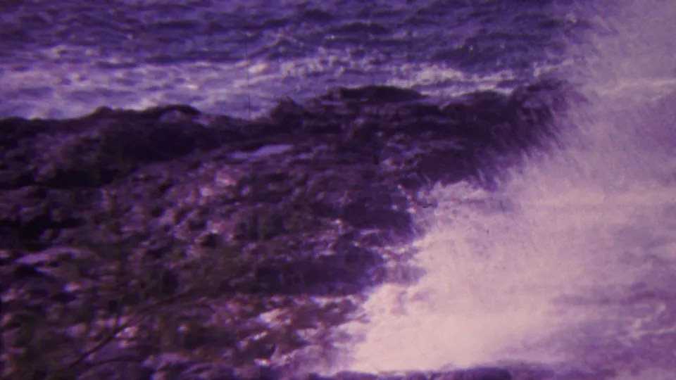 1962 Hawaii 8mm frame showing white ocean spray bursting against dark shoreline rocks with blurred low vegetation at the edge of the frame.
