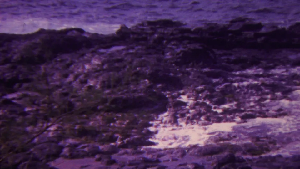 1962 Hawaii 8mm frame of dark coastal rocks, white foam pooling in channels, and rough ocean water along the shoreline.