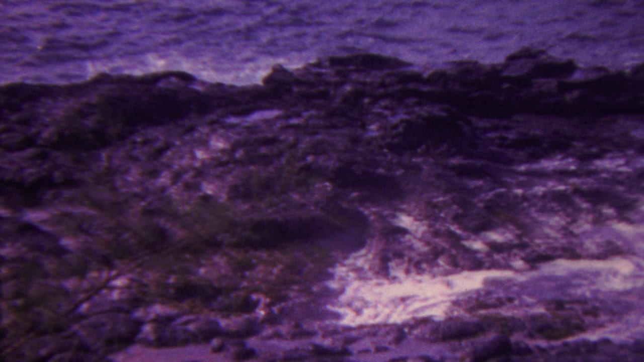 1962 Hawaii 8mm frame showing rough ocean water washing around dark volcanic-looking shore rocks with white foam and a purple aged-film color shift.
