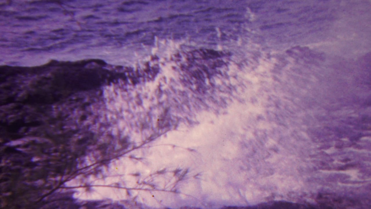1962 Hawaii 8mm frame with a wave striking jagged shoreline rocks, sending pale spray upward while a thin foreground branch crosses the scene.
