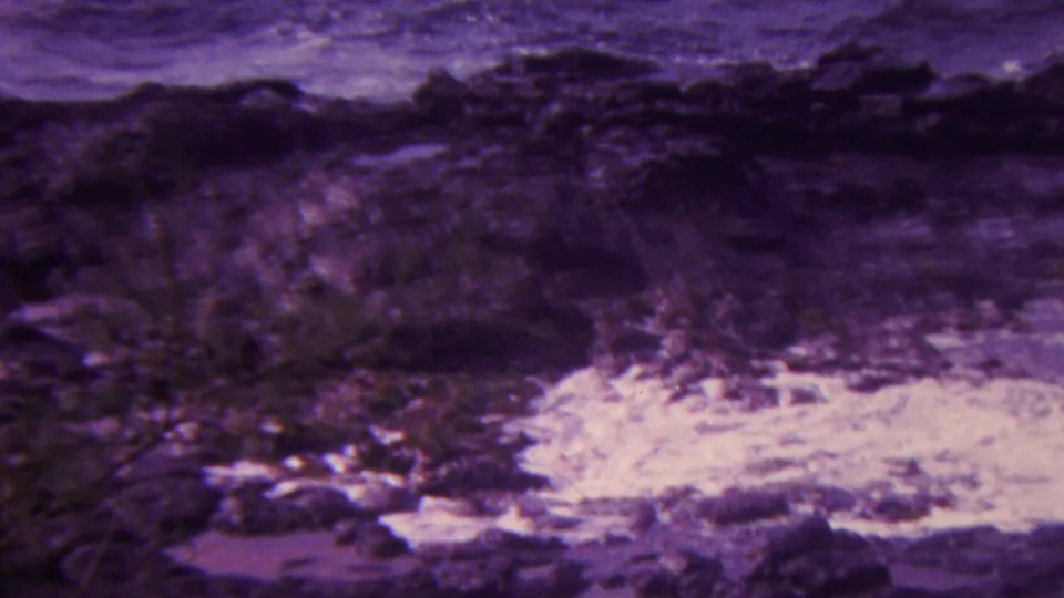1962 Hawaii 8mm frame showing white foam pooling and moving around dark coastal rocks, with soft focus and vegetation near the lower edge.