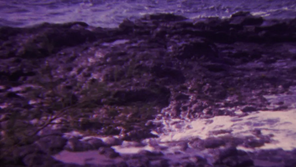 1962 Hawaii 8mm frame of white surf breaking across dark, uneven shoreline rocks with rough ocean water beyond and strong purple color fading.