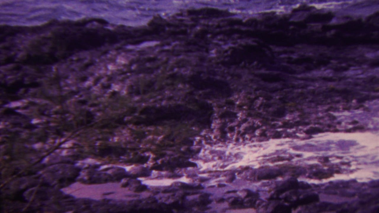 1962 Hawaii home-movie frame showing foamy ocean water moving through dark rocky shoreline formations with a purple film cast.
