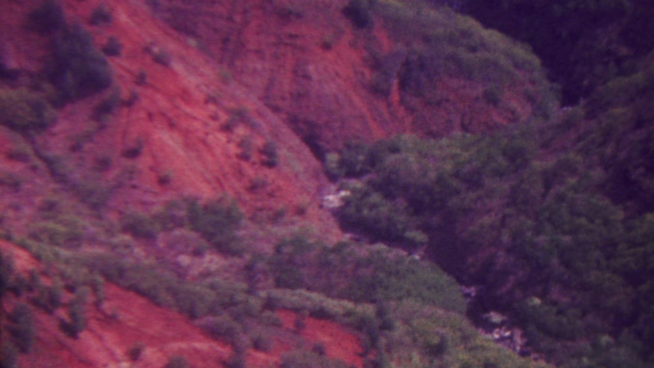 1962 Hawaii 8mm frame showing a color-shifted rugged hillside, patches of green vegetation, and a narrow ravine cutting through the terrain.