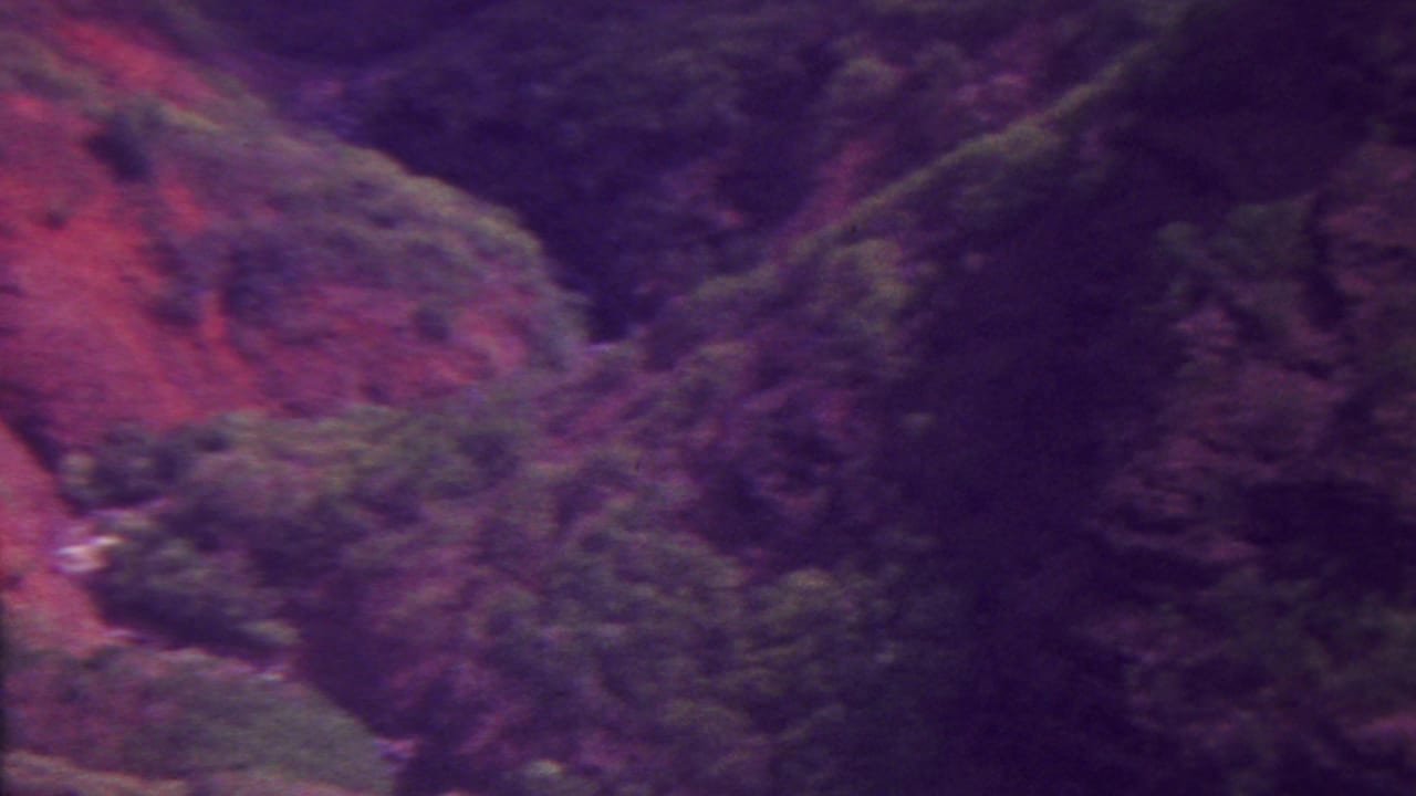1962 Hawaii 8mm frame with a steep red-toned slope, dark valley shadow, and clusters of green vegetation in a rugged landscape.