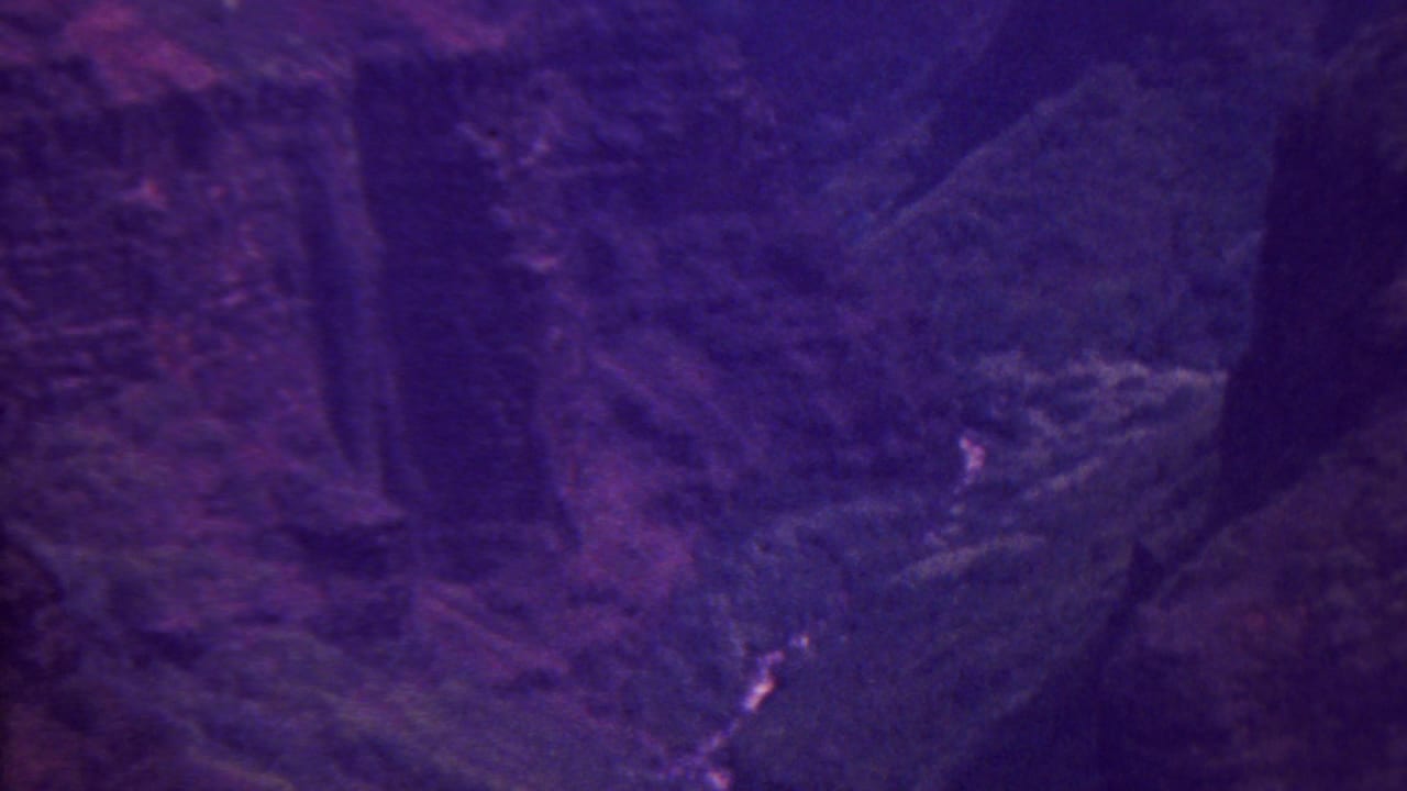 1962 Hawaii 8mm frame showing a high view over steep canyon terrain, forested slopes, and a thin winding stream under a strong purple-magenta film cast.