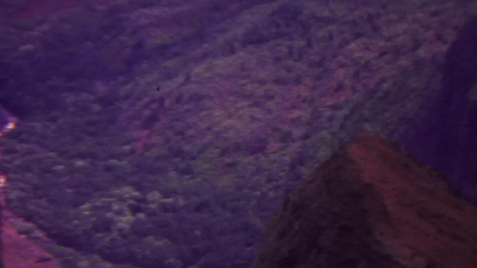 1962 Hawaii 8mm frame showing an aerial view over rugged, forested mountain terrain with reddish exposed slopes and strong purple color shift.