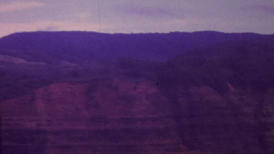 1962 Hawaii 8mm frame showing a distant mountain ridge and hazy valley floor, with reddish terrain and soft purple-blue film coloration.