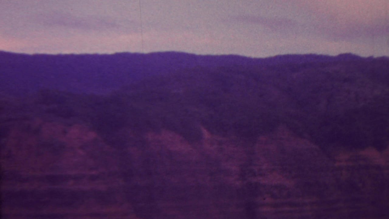 A 1962 Hawaii travel-film frame, per source metadata, showing a dark ridge and reddish canyon wall beneath a soft purple-pink dusk sky.