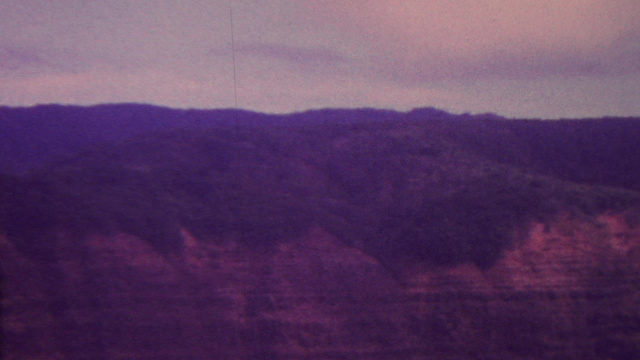 A source-attributed 1962 Hawaii 8mm frame showing layered reddish rock formations, dark upper slopes, and a hazy evening sky.