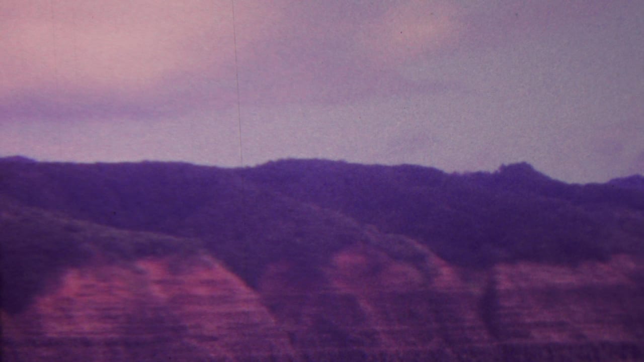 1962 Hawaii 8mm frame showing distant mountain ridges, layered cliff faces, and a cloudy sky with strong magenta-blue color fading.