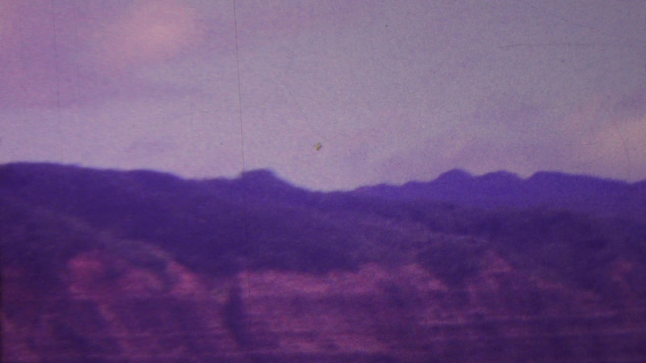 1962 Hawaii home-movie frame with a dark ridge line, reddish cliff slopes, soft cloud cover, and visible 8mm grain and scratches.