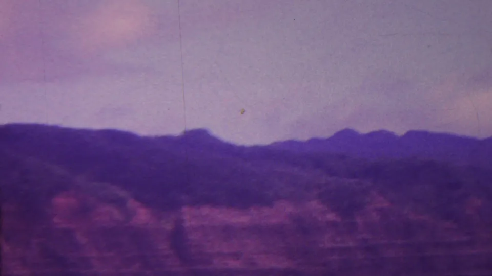 1962 Hawaii home-movie frame with a dark ridge line, reddish cliff slopes, soft cloud cover, and visible 8mm grain and scratches.