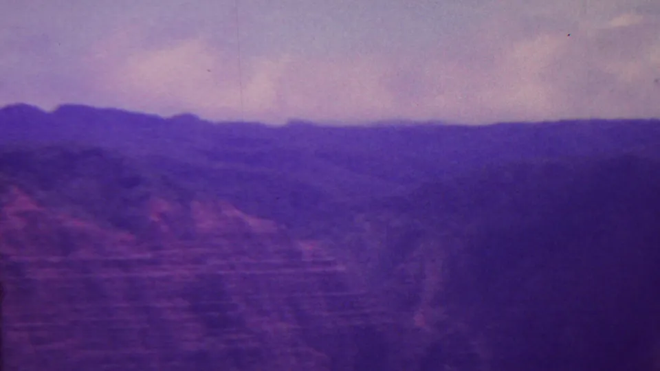 1962 Hawaii 8mm frame showing a shadowed cliff face, valley forms, distant blue ridges, and a cloudy sky with heavy color shift.