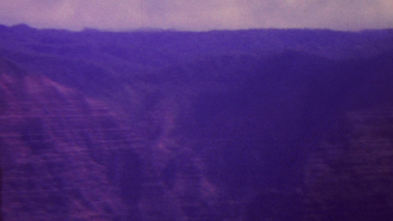 1962 Hawaii 8mm frame showing distant mountain ridges, dark canyon slopes, and a soft overcast sky with strong purple film fading.