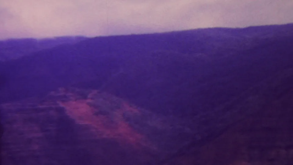 1962 Hawaii 8mm frame looking across rugged canyon terrain, with layered hills, vegetation texture, and a strong magenta-purple archival cast.
