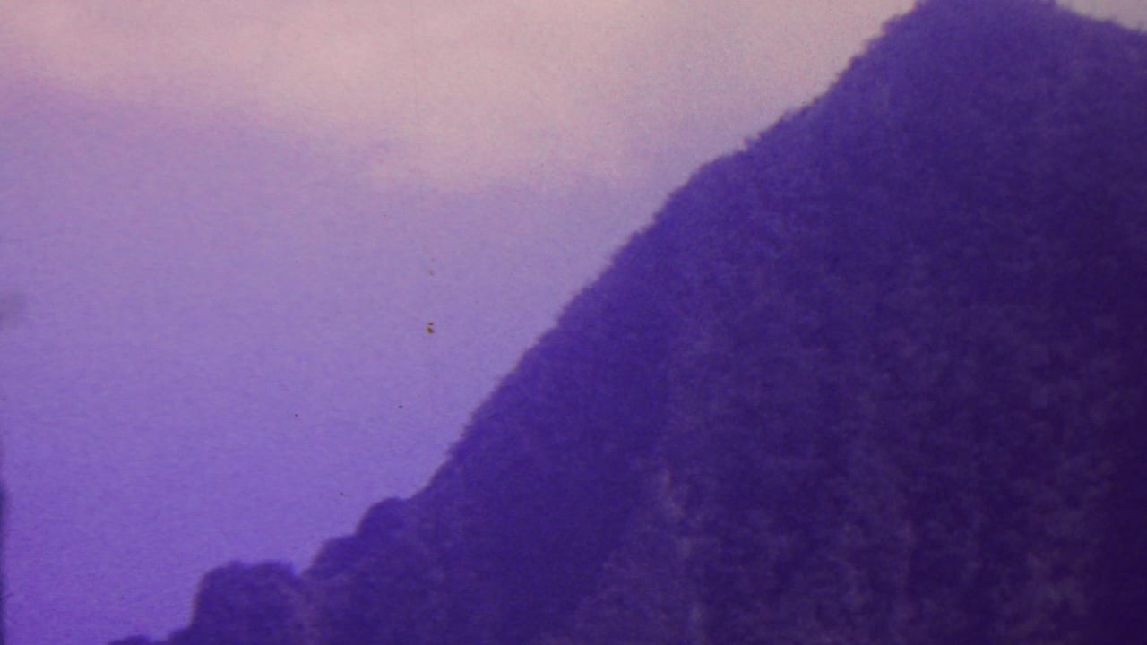 1962 Hawaii home-movie frame with rugged, tree-covered mountain forms on both sides of a pale purple sky opening.