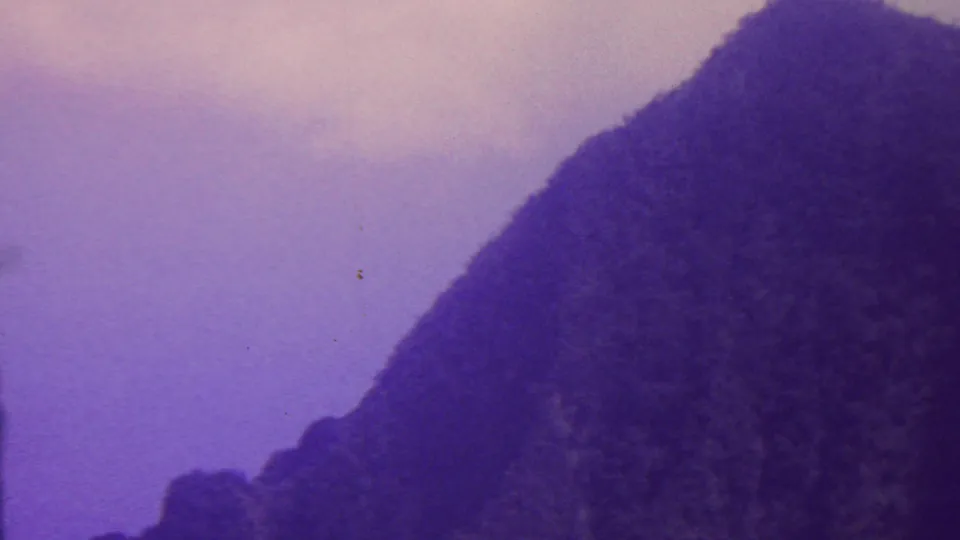 1962 Hawaii home-movie frame with rugged, tree-covered mountain forms on both sides of a pale purple sky opening.