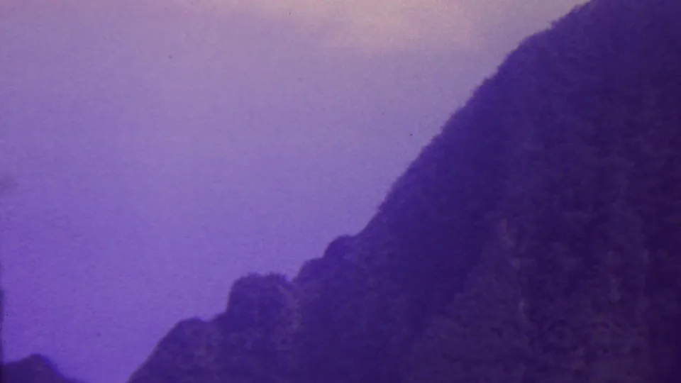 1962 Hawaii 8mm frame showing a steep dark mountain face with lower rocky shapes and a lavender sky, rendered with grain and muted color shift.