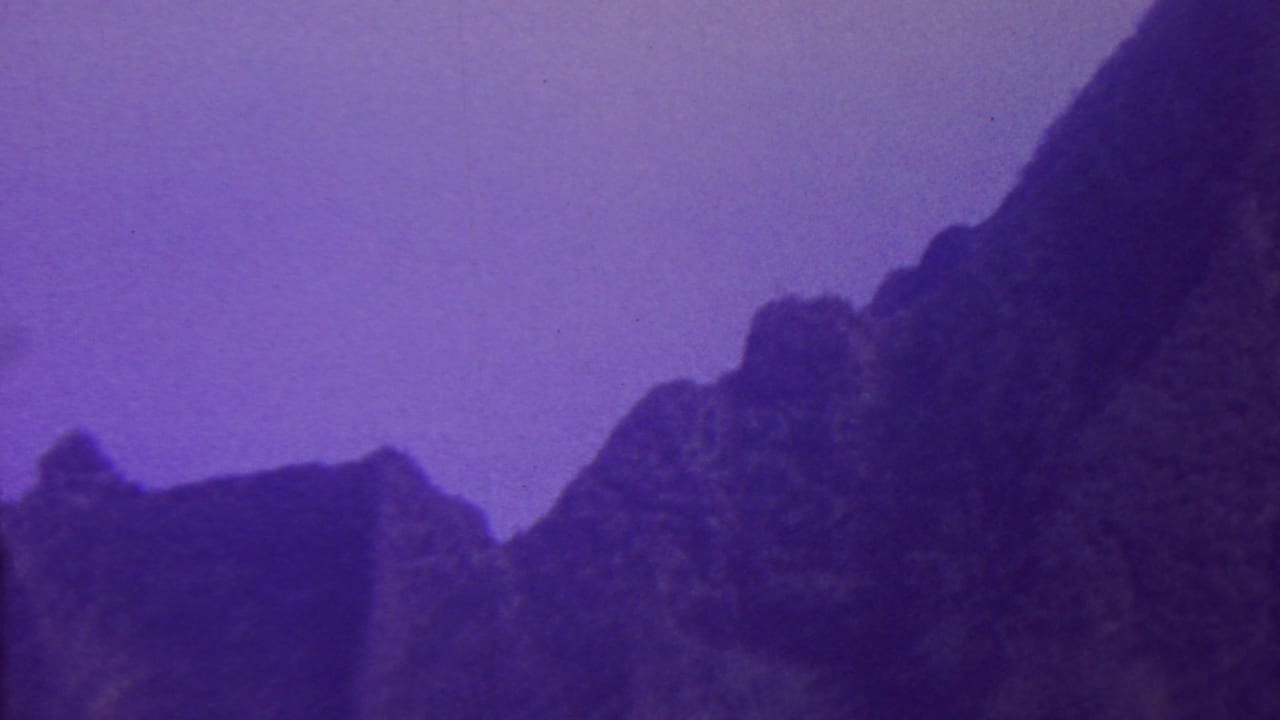1962 Hawaii 8mm frame showing a dark, craggy mountain ridge rising into a purple-blue sky, with soft grain and aged color shift.