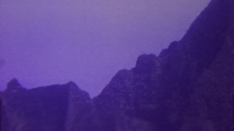 1962 Hawaii 8mm frame showing a dark, craggy mountain ridge rising into a purple-blue sky, with soft grain and aged color shift.