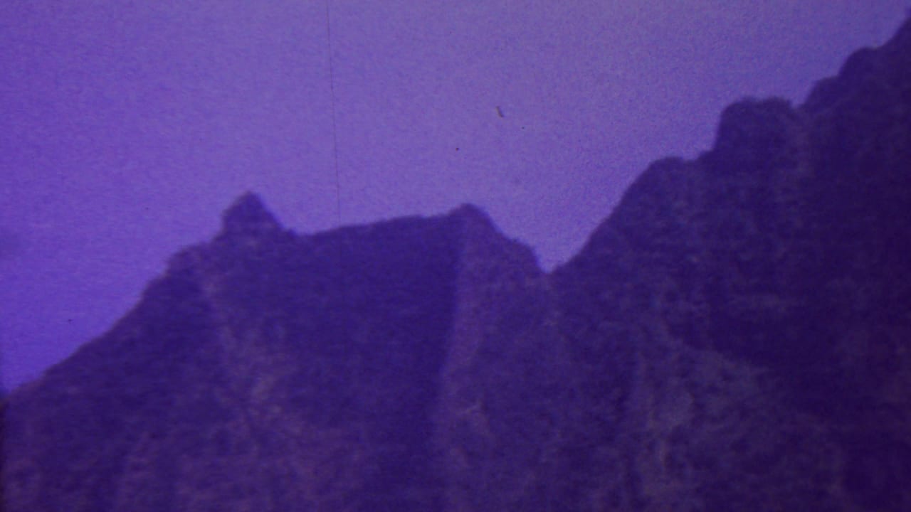 1962 Hawaii 8mm frame with rugged mountain peaks and shadowed ridge forms under a purple sky, recorded with soft focus and visible film grain.