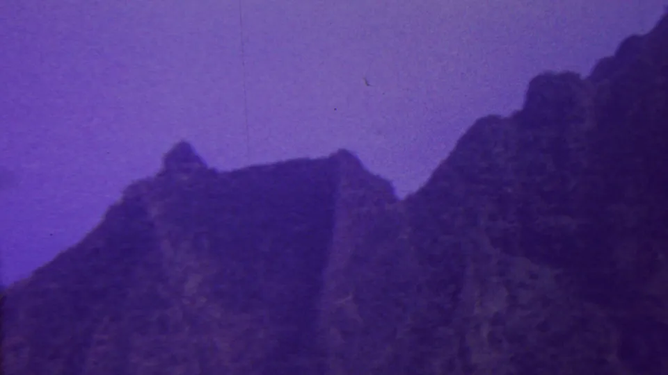 1962 Hawaii 8mm frame with rugged mountain peaks and shadowed ridge forms under a purple sky, recorded with soft focus and visible film grain.