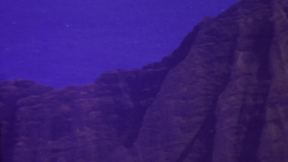 1962 Hawaii 8mm frame showing a shadowed rocky mountain slope rising into a blue-purple sky, with soft focus and visible film grain.