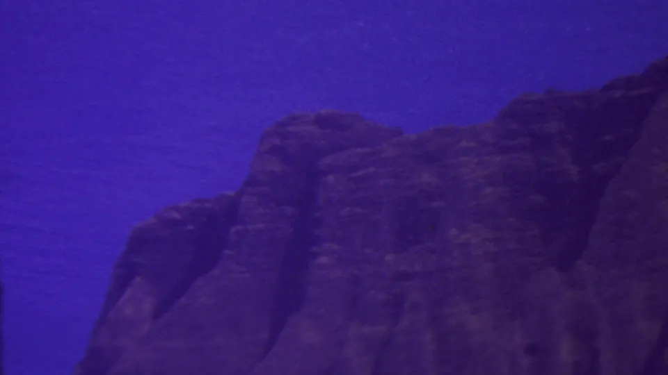1962 Hawaii 8mm frame showing a broad dark mountain ridge and cliff shapes under a saturated blue-purple sky, with no people or structures visible.