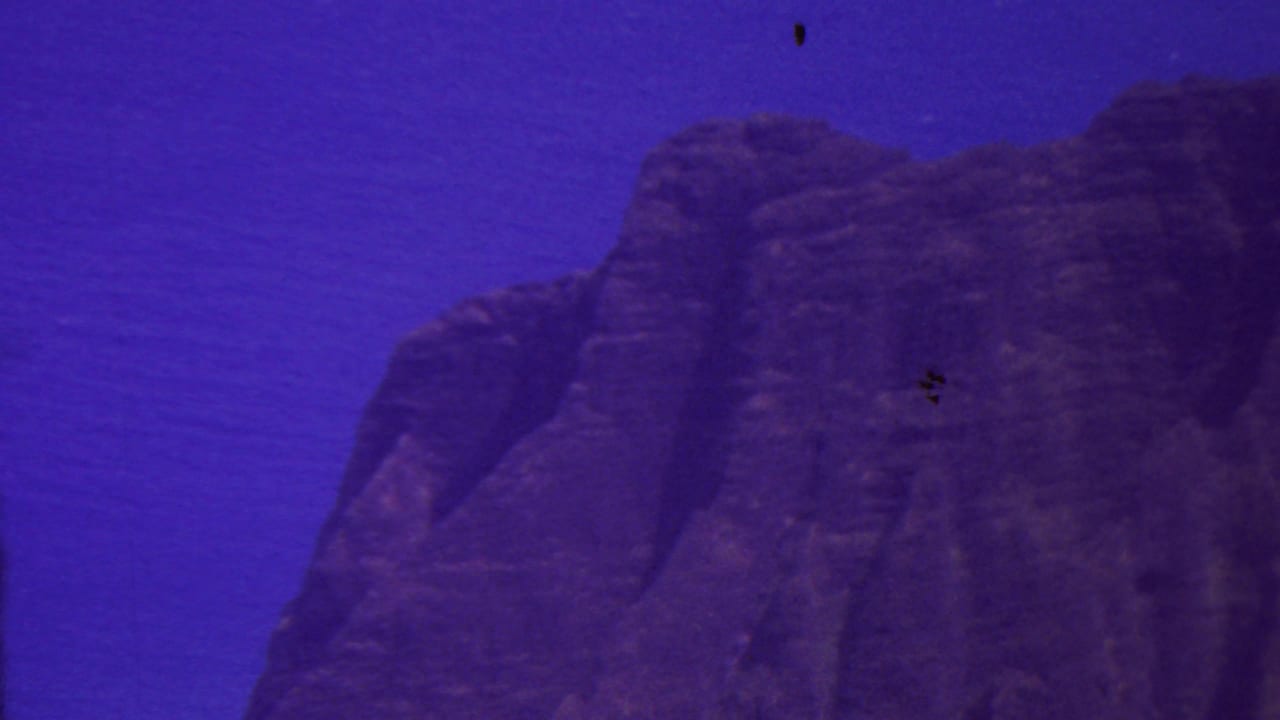 1962 Hawaii 8mm frame showing a steep dark coastal cliff rising beside deep blue water under an open sky, with aged-film purple-blue color shift.