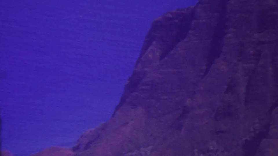 1962 Hawaii 8mm frame looking across a high rocky escarpment, with the cliff filling the right side and blue ocean or distant water behind it.